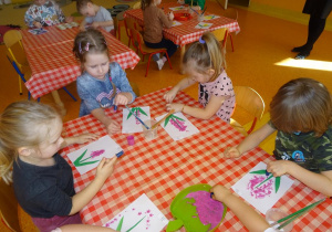 Czwórka dzieci stempluje paluszkami płatki kwiatów na kartce papieru wokół zielonej łogygi.